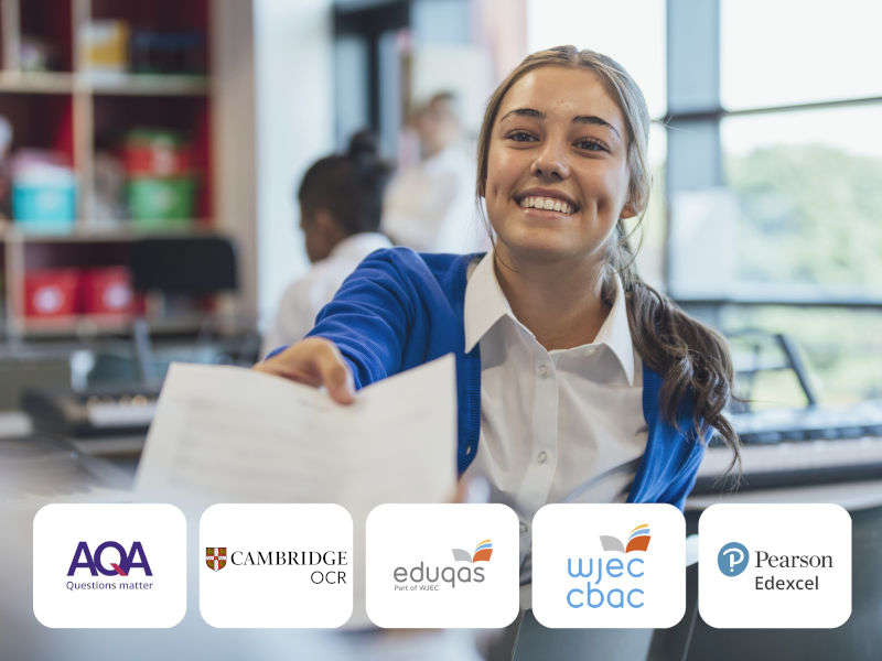 Smiling student in school uniform extending worksheet toward viewer in bright classroom, with exam board logos (AQA, OCR, Eduqas, WJEC CBAC, Pearson Edexcel) displayed below